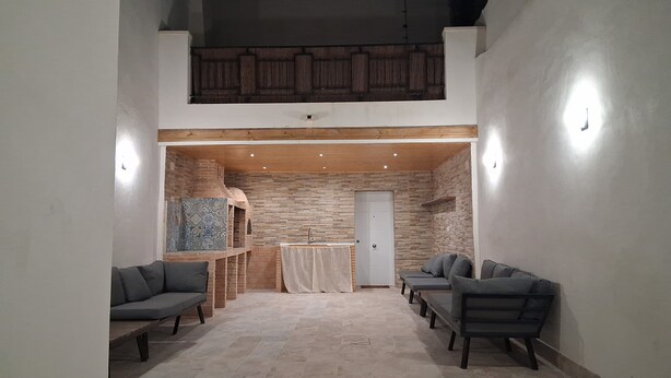 Una habitación amplia con techo alto, chimenea empotrada y pared de piedra. Hay dos sofás y un banco.