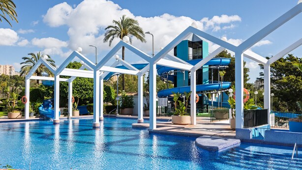 A swimming pool with a water slide, palm trees, and a clear blue sky.
