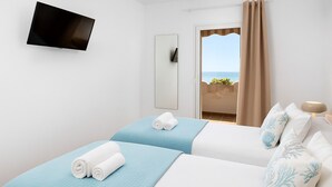 A hotel room with two beds, a TV mounted on the wall, a balcony with a view of the sea, and a door leading to an outdoor area.