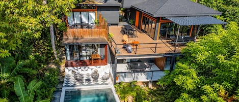 Una casa moderna de dos pisos con una terraza en la azotea, piscina y exuberante vegetación alrededor de la propiedad.