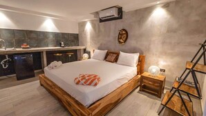 Een slaapkamer met een houten bed, een klein nachtkastje met een lamp, een wandairconditioner en een keukenhoek met een spoelbak en krukken.
