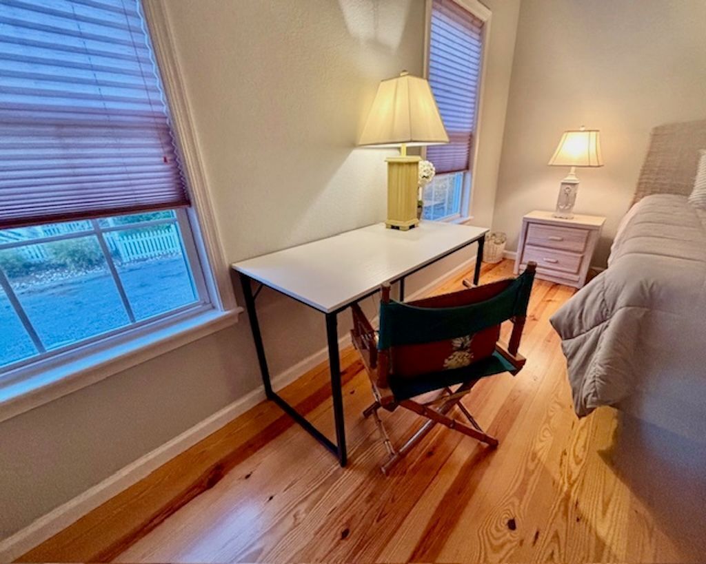 A bedroom with a bed, a desk, and a chair. There are two windows with blinds, a nightstand with a lamp, and a view of the outside.