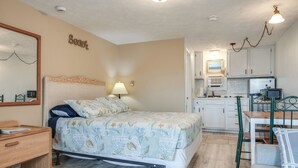 Een slaapkamer met een bed, nachtkastje en een klein bureau. Er is een kitchenette met een koelkast, kookplaat en spoelbak.