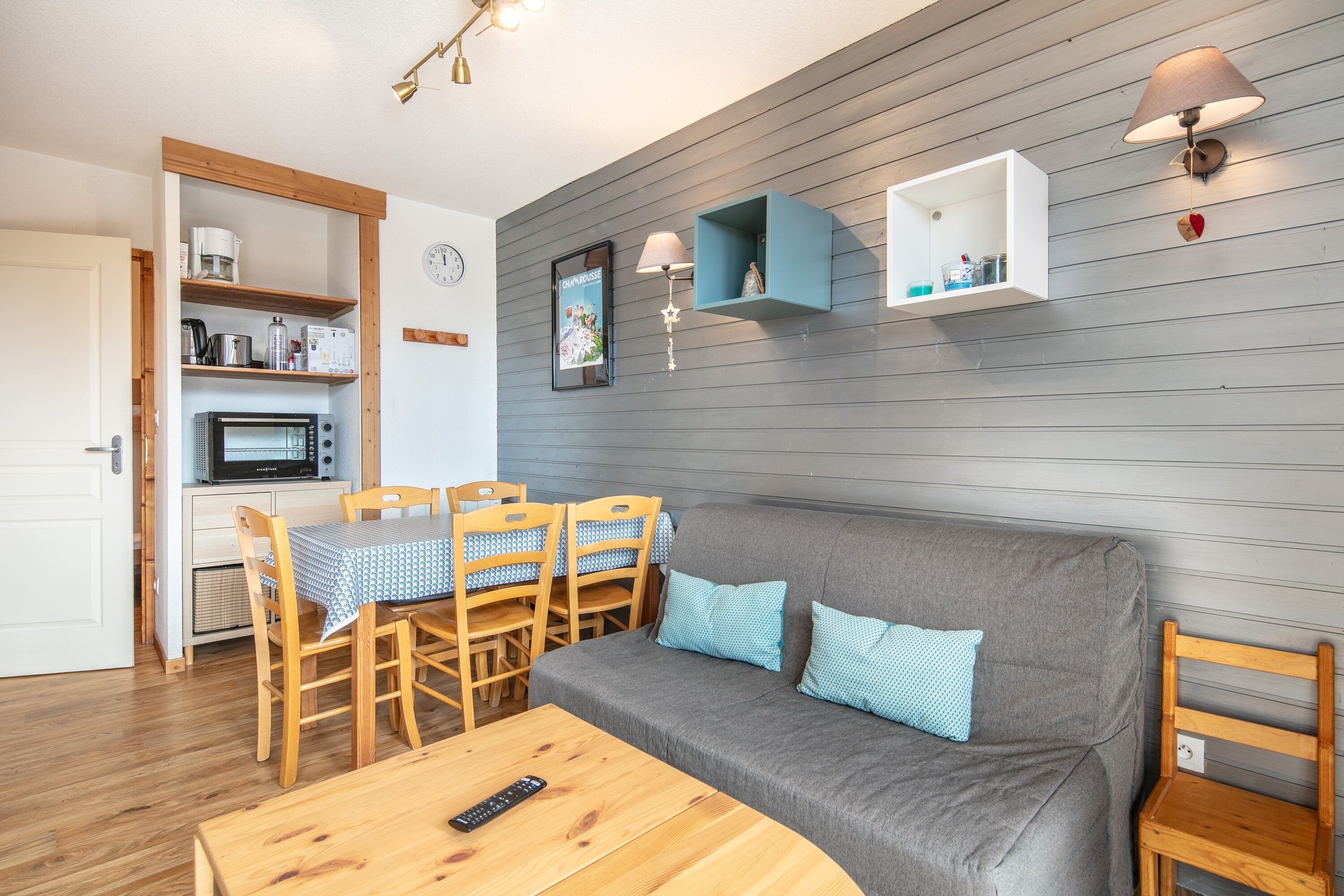 A cozy living room with a grey sofa, wooden dining table, and chairs. There is a microwave and a clock on the wall.