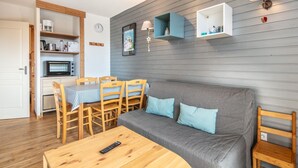 Un salon chaleureux avec un canapé gris, une table à manger en bois et des chaises. On y trouve également un four à micro-ondes et une horloge murale.
