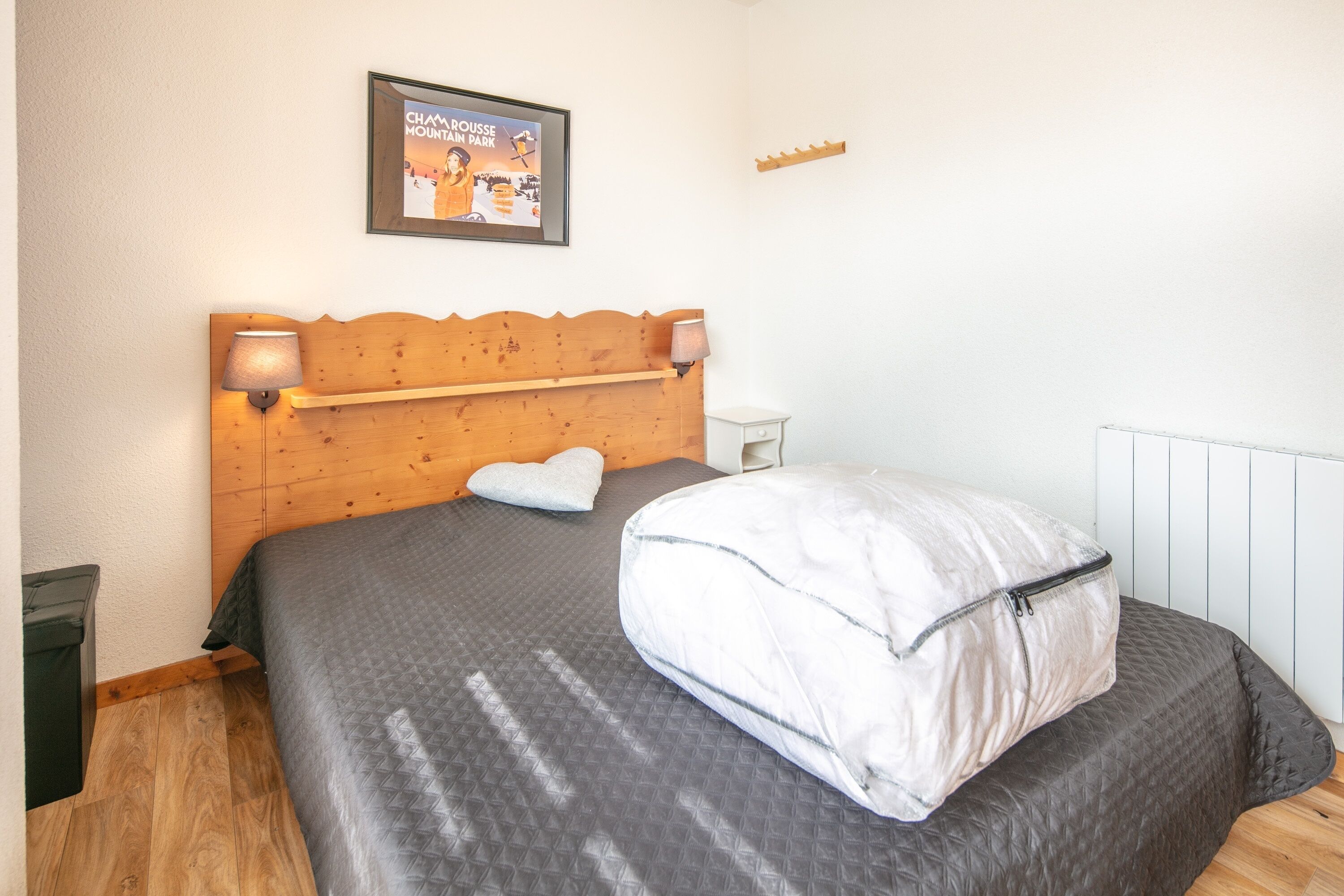 Ein Schlafzimmer mit einem hölzernen Kopfteil, einem Bett mit grauer Tagesdecke, einem weißen Kissen, einem gerahmten Bild an der Wand und einem kleinen Nachttisch.