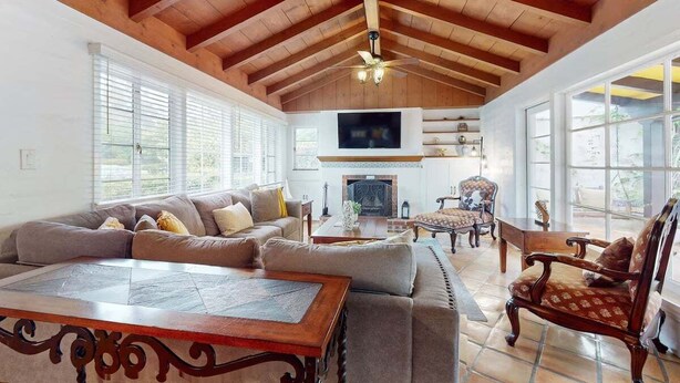 A spacious living room with a high ceiling, wooden beams, a fireplace, and large windows. - La Jolla Historic Gem | Hot Tub, Game Room, Garden Oasis w/ Firepit & Courtyard Dining (La Jolla)