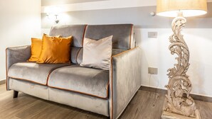 Een kamer met een bakstenen plafond, een grijze bank met oranje kussens, een witte lamp en een houten vloer.
