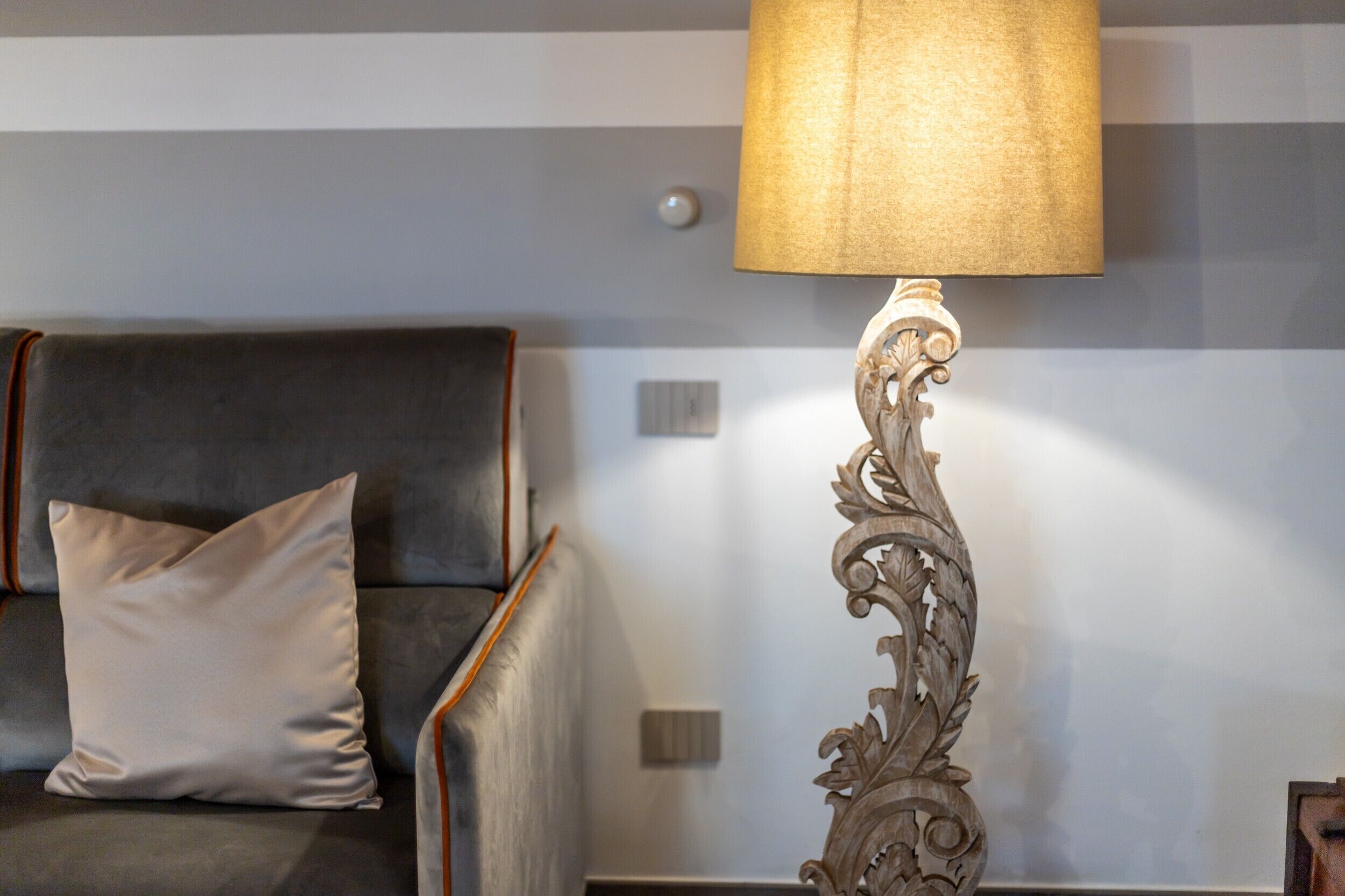 A sofa with a cushion, a wall with a light switch, and a decorative floor lamp.