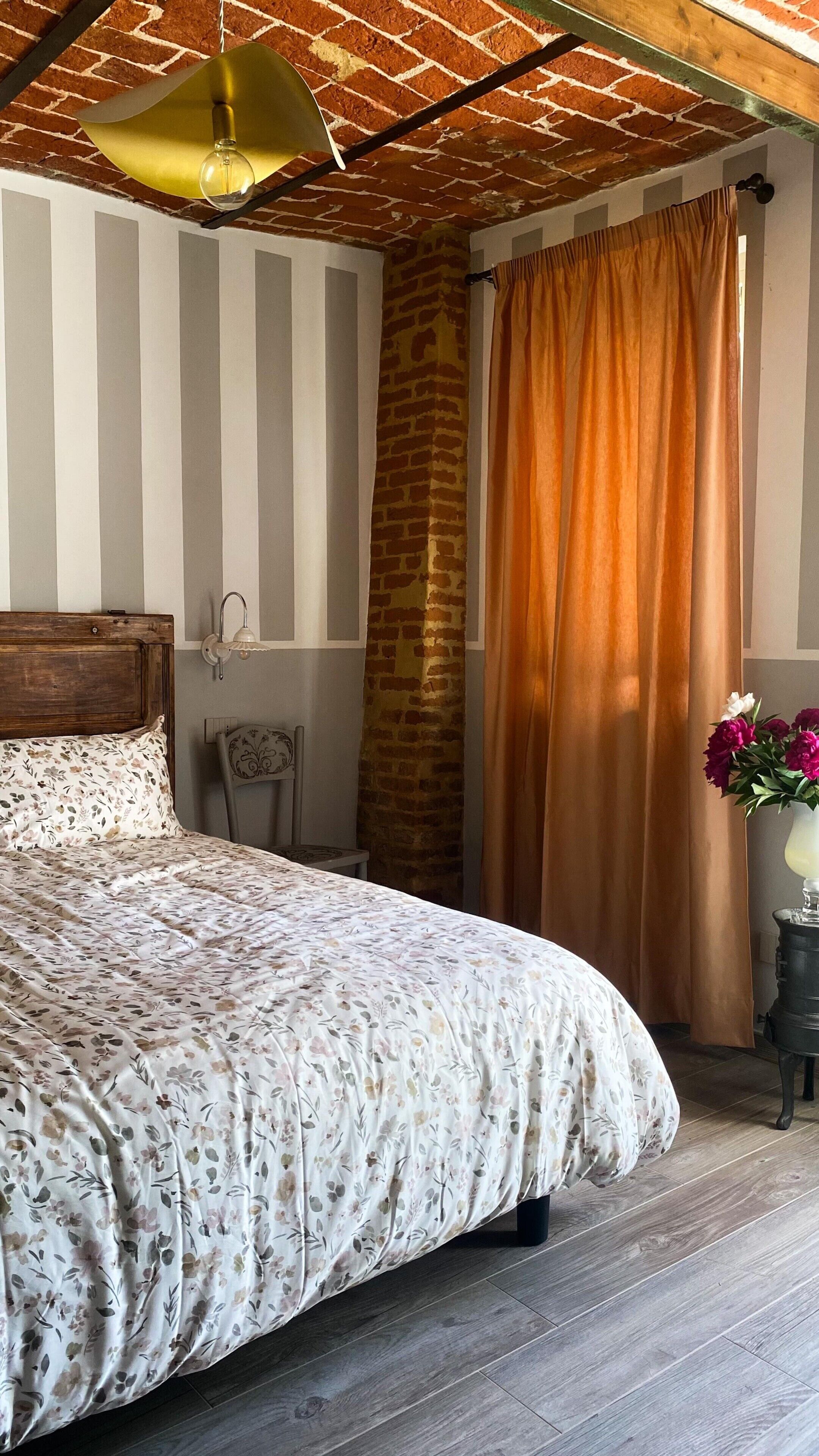 A bedroom with a bed, a nightstand, a lamp, a brick wall, and a window with curtains.