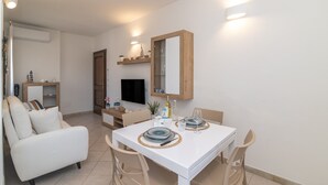 A modern living room with a white sofa, a dining table set with plates and glasses, a TV mounted on the wall, and a wooden cabinet with decorative items.