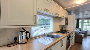 Eine moderne Küche mit weißen Schränken, einer Edelstahlspüle und einem Wasserkocher. Ein Fenster mit Jalousien, ein Herd und ein Essbereich mit einer Couch sind ebenfalls vorhanden.