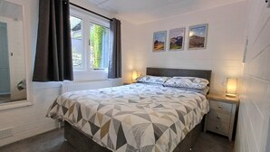 Ein Schlafzimmer mit einem Bett, zwei Nachttischen, einem Fenster mit Vorhängen, einer Heizung und drei gerahmten Bildern an der Wand.