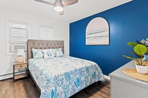 Ein Schlafzimmer mit einem karottenförmigen Kopfteil, einem Bett mit floralem Überwurf, einem Deckenventilator, einem Wandbild und einem Topf mit Pflanze.