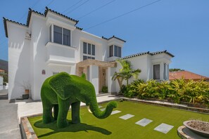 Un bâtiment blanc avec un toit recouvert de tuiles, une pelouse bien entretenue et une grande sculpture d’éléphant verte.
