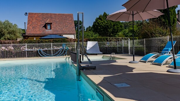Una piscina amb gandules i ombrel·les, envoltada per una tanca i un edifici amb teulada de rajola.