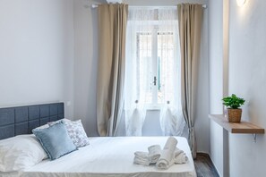 A neatly made bed with white linens and rolled towels, a grey upholstered headboard, and a potted plant on a wooden shelf.