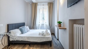 Un dormitorio con una cama, una mesita de noche, un televisor montado en la pared y una ventana con cortinas.