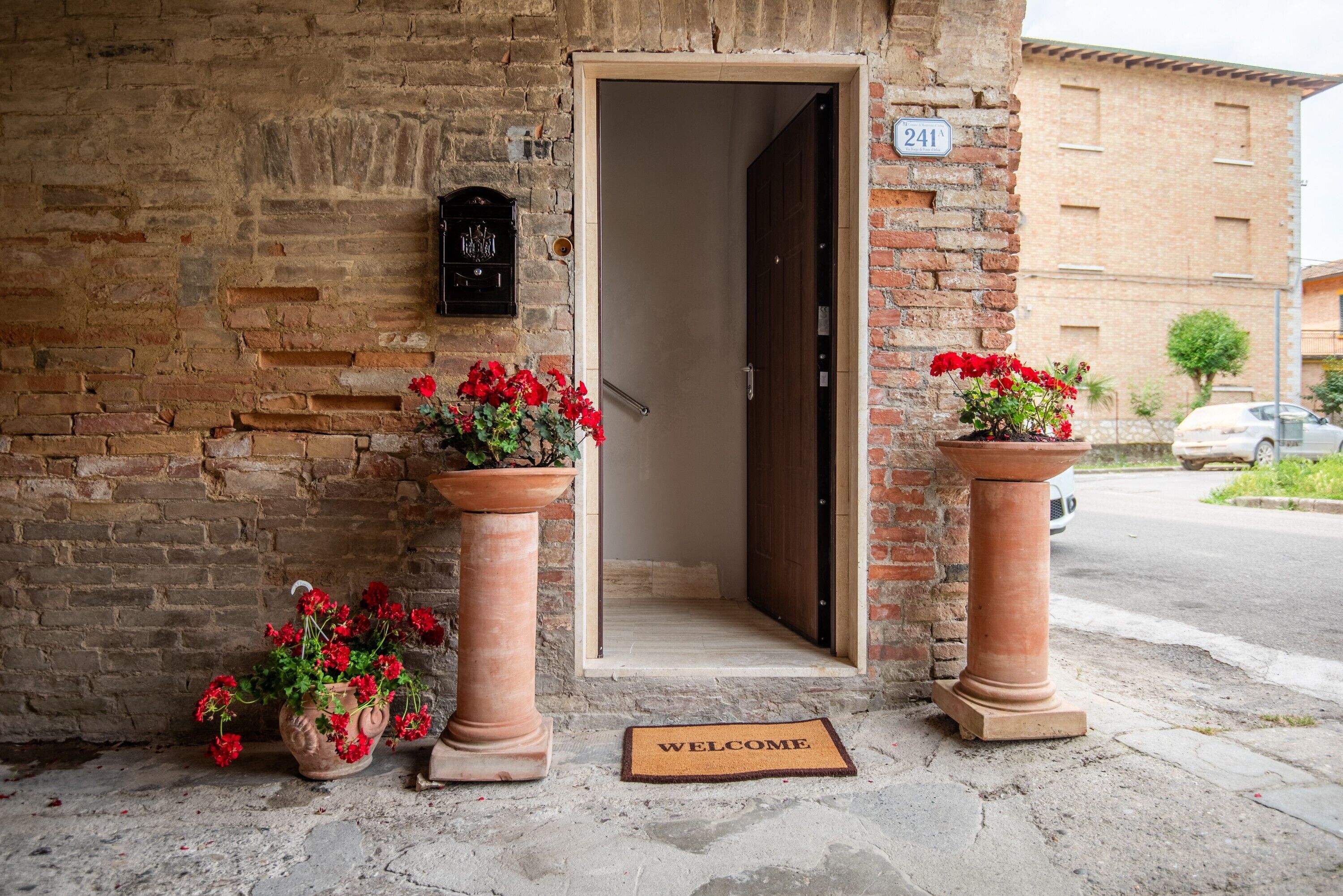 Um edifício de tijolos com porta, o número 241, e dois vasos com plantas de flores vermelhas.