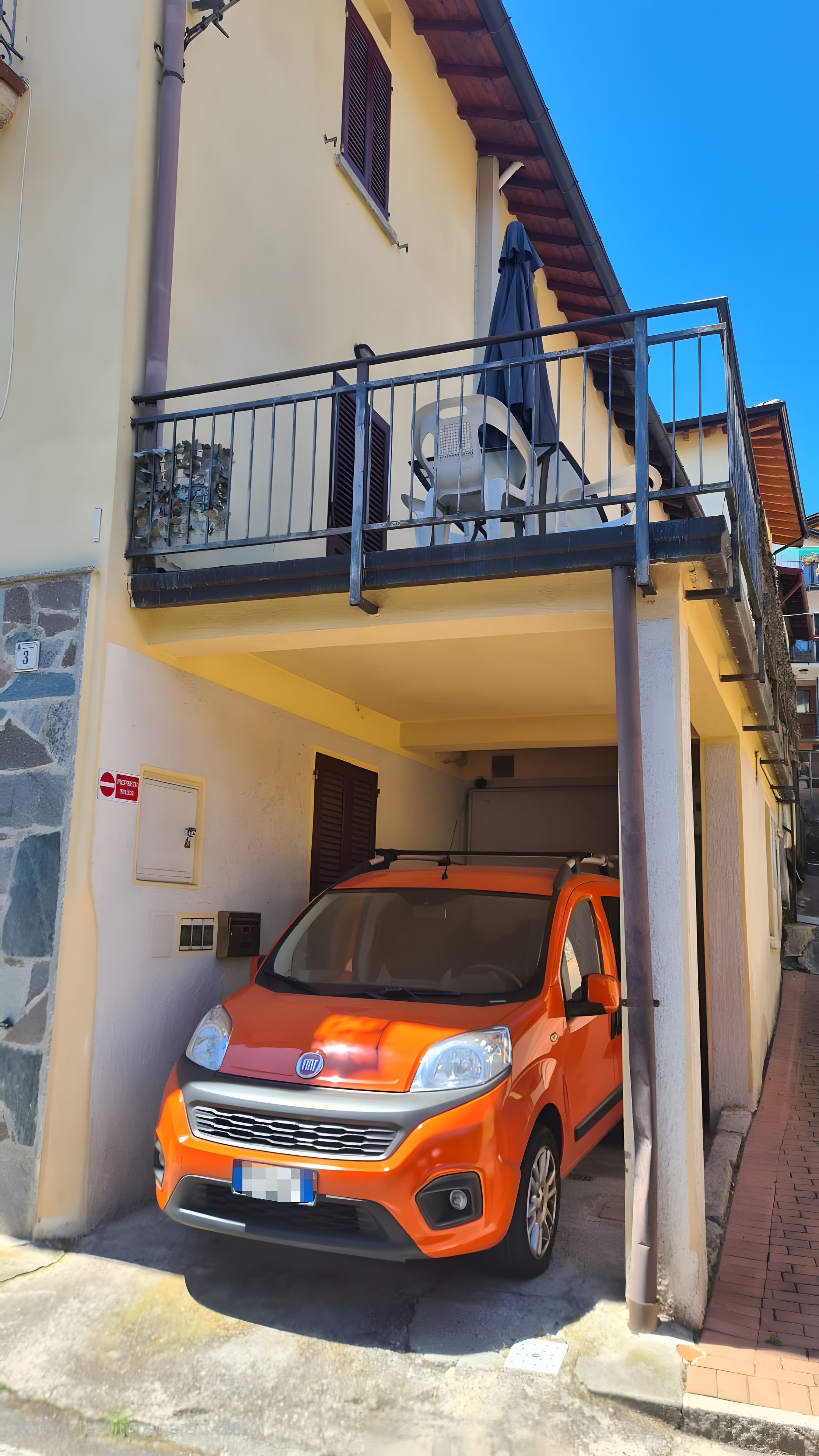 Une voiture orange est garée devant un immeuble doté d’un balcon, d’un mur en pierre et d’un panneau indiquant « accès interdit ».
