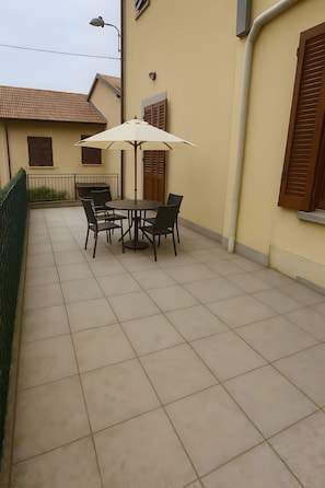 Une terrasse extérieure avec une table ronde et quatre chaises, un parasol blanc et un lampadaire.