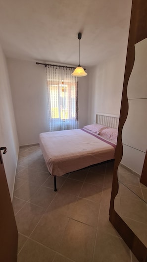 A bedroom with a bed, a window with curtains, a hanging light fixture, and a radiator.