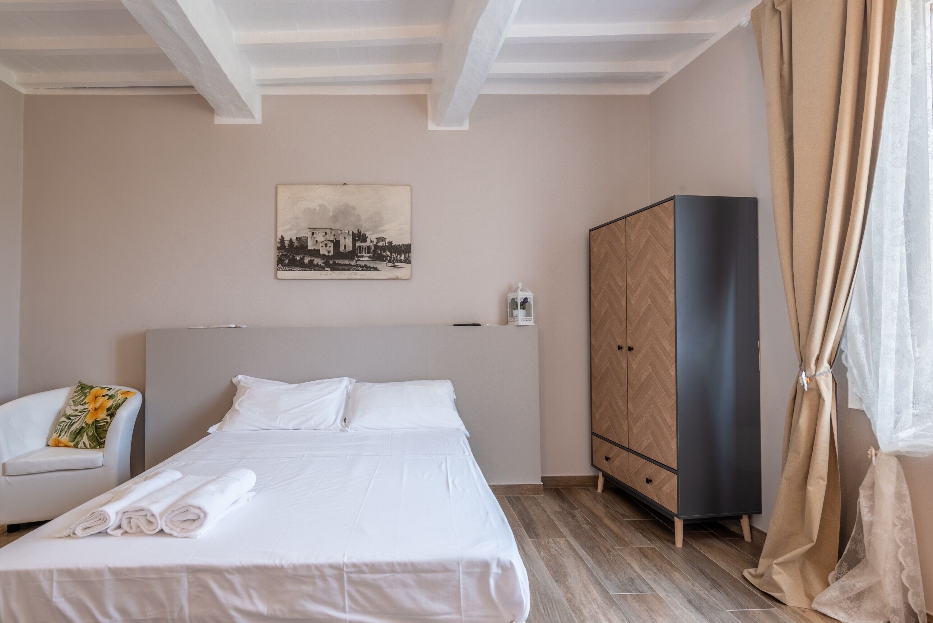 Ein ordentlich bezogenes Bett mit weißer Bettwäsche, ein Stuhl mit einem floralen Kissen, ein Kleiderschrank und ein an der Wand hängendes Bild.