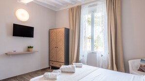 A hotel room with a bed, a TV mounted on the wall, a wardrobe, a window with curtains, and a small shelf with a plant.