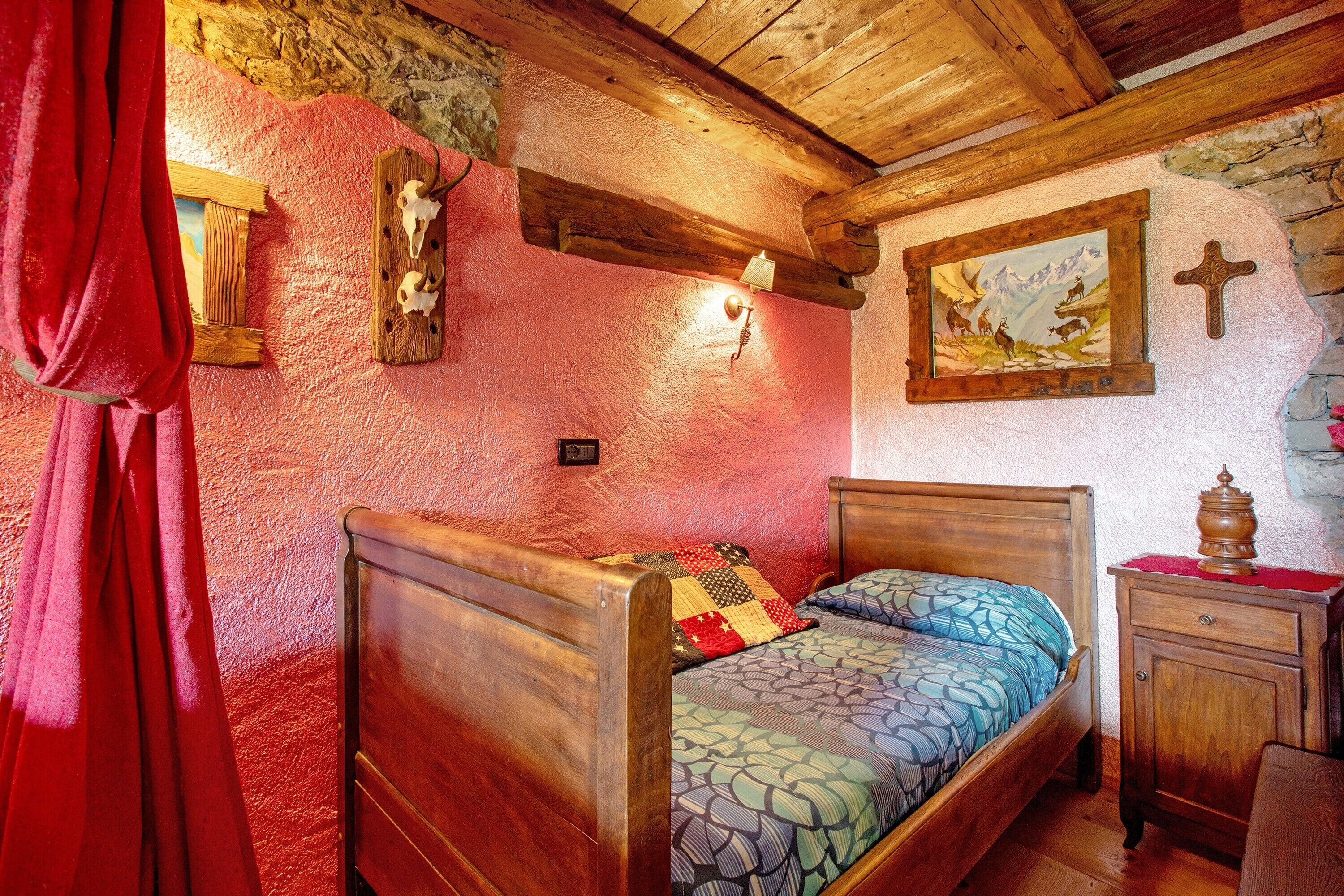 Una cama de madera con colcha estampada, una mesita de noche de madera, una lámpara fijada a la pared y un cuadro enmarcado con montañas.