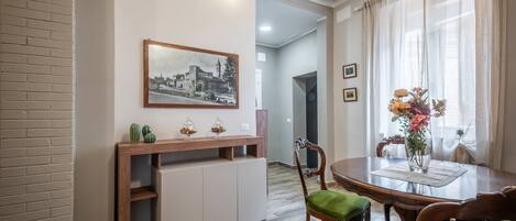 A dining area with a wooden table, chairs, a vase with flowers, a framed picture on the wall, and a brick wall.