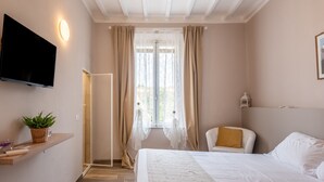 Un dormitorio con cama, televisor montado en la pared, planta sobre una estantería, una silla y ventana con cortinas.