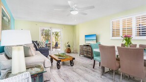 A living room with a ceiling fan, a TV, a dining area with chairs, and a view of the outdoors through a sliding door. - Lulu's Crescent Cottages - Entire two Unit Complex (Siesta Key)