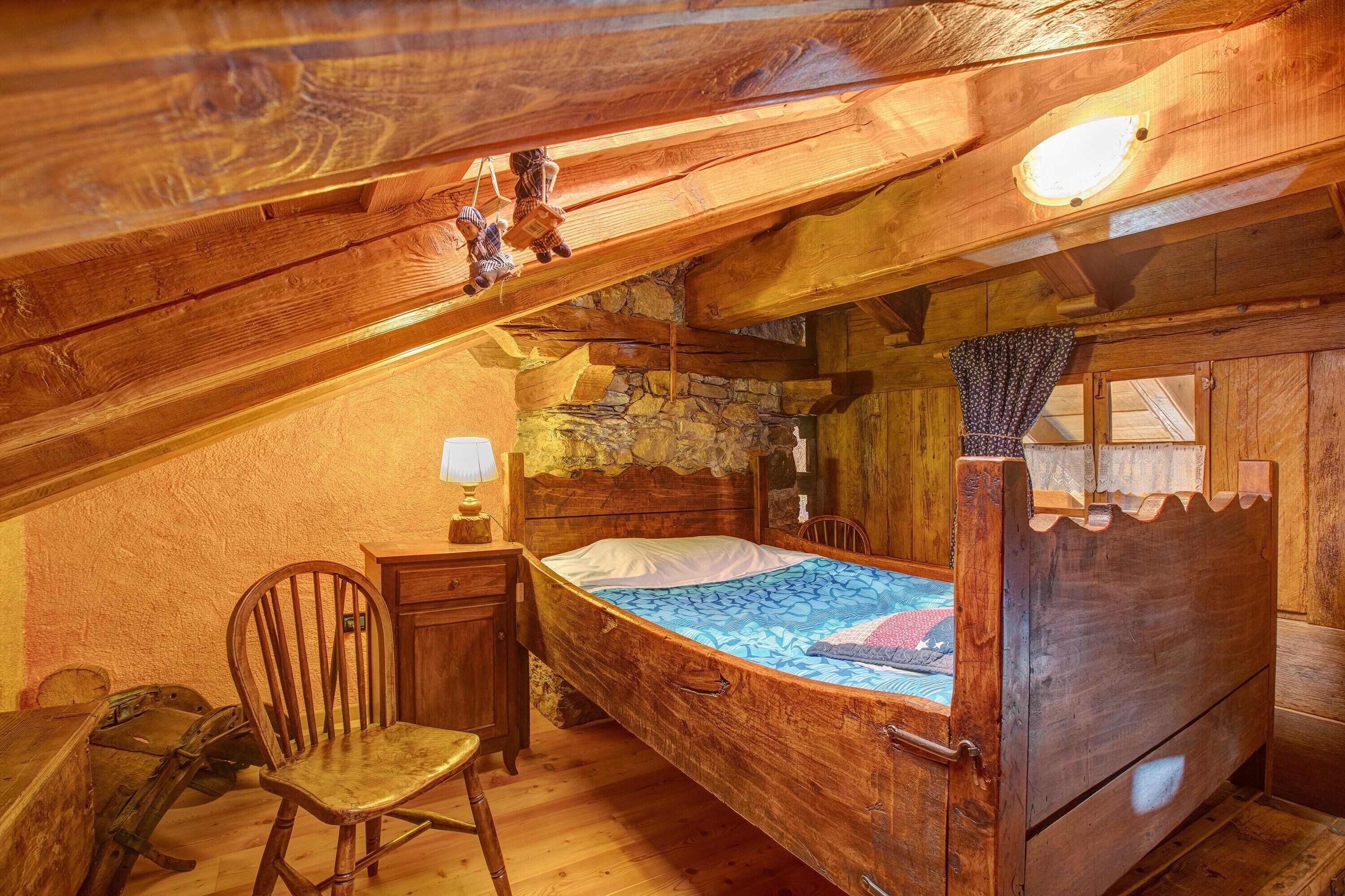 A wooden room with a bed, a chair, a desk, and a stone wall.