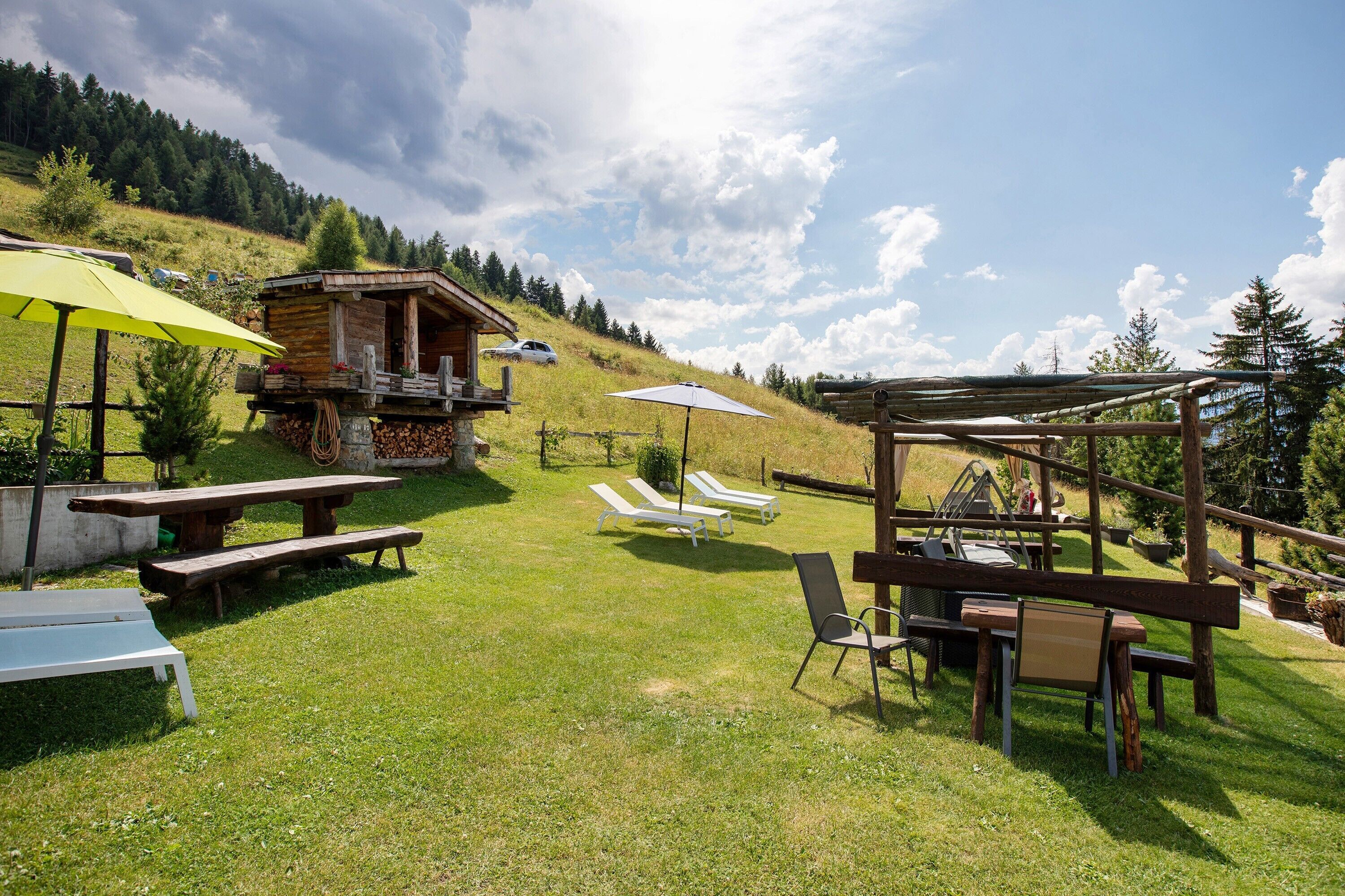 A rustic outdoor setting with wooden benches, a barbecue area, and a small cabin on a grassy hill.