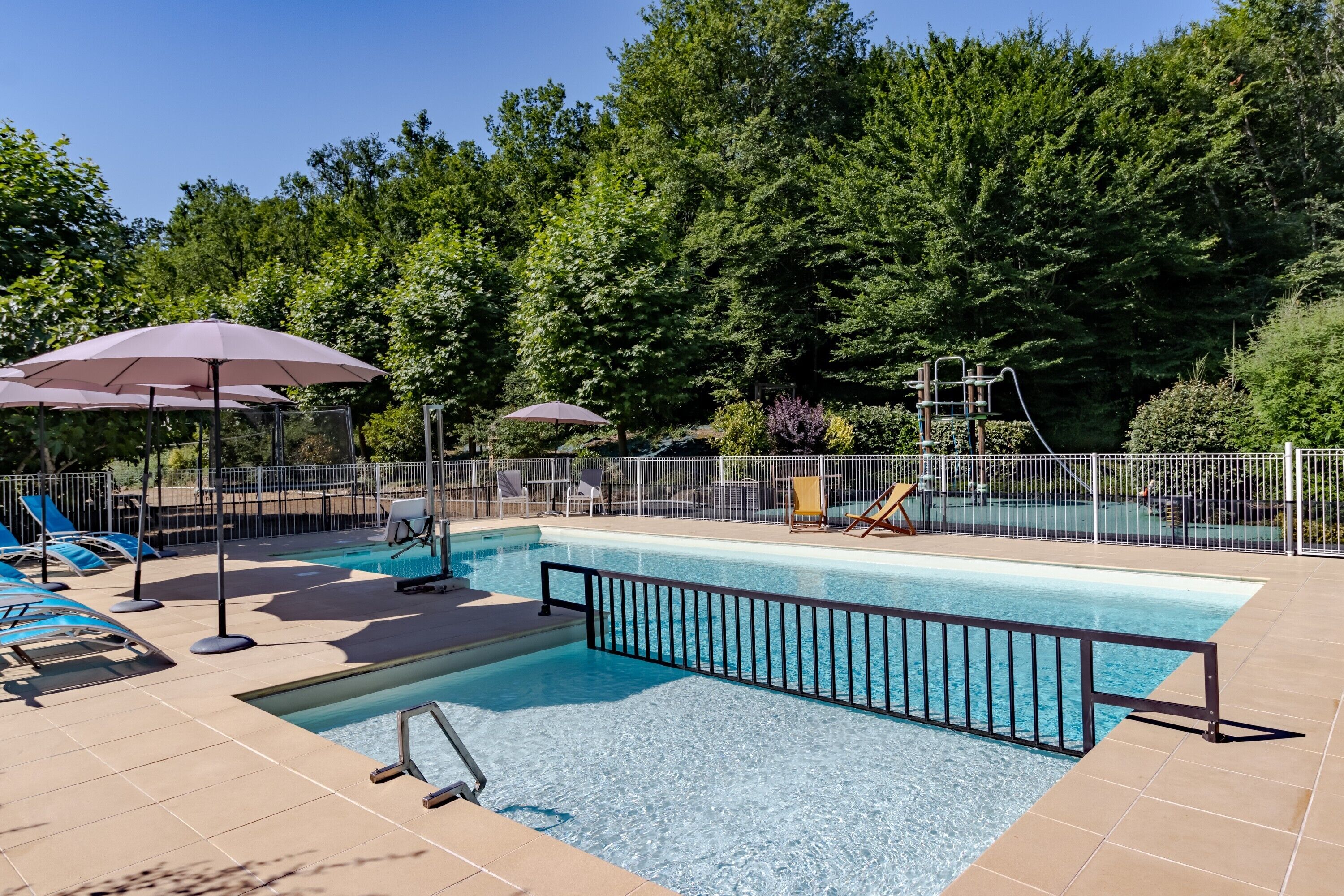 Piscina com espreguiçadeiras e guarda-sóis, rodeada por uma área cercada e árvores.