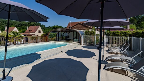 Ein Schwimmbecken mit Liegestühlen und Sonnenschirmen, umgeben von einem Zaun, mit Häusern im Hintergrund.
