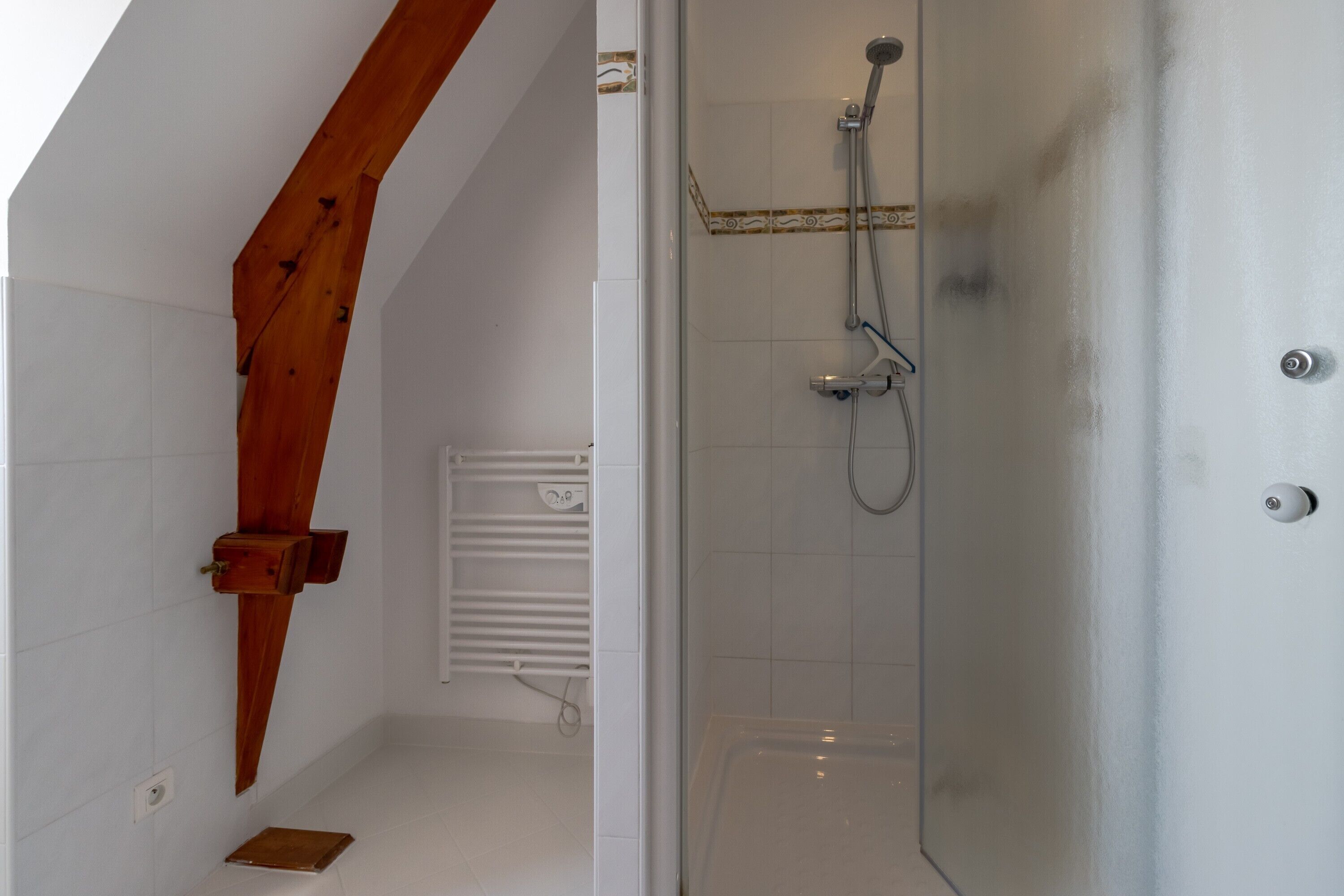 A shower with a glass door, a wooden beam, and a white tiled wall.