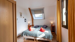 Ein Schlafzimmer mit schrägem Dach, einem Oberlicht, einem Bett mit gesteppter blauer Bettdecke, zwei Kissen, einem Nachttisch mit Lampe und einer Holzleiter.
