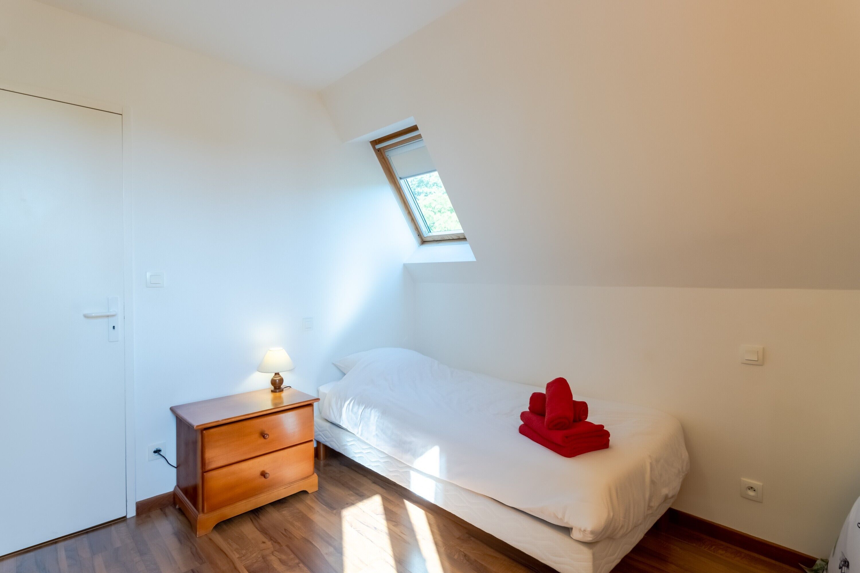 Una habitación pequeña y luminosa con una cama individual, una mesita de noche, una lámpara y un tragaluz.