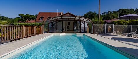 A swimming pool with a covered area, surrounded by a fence, and a house in the background.