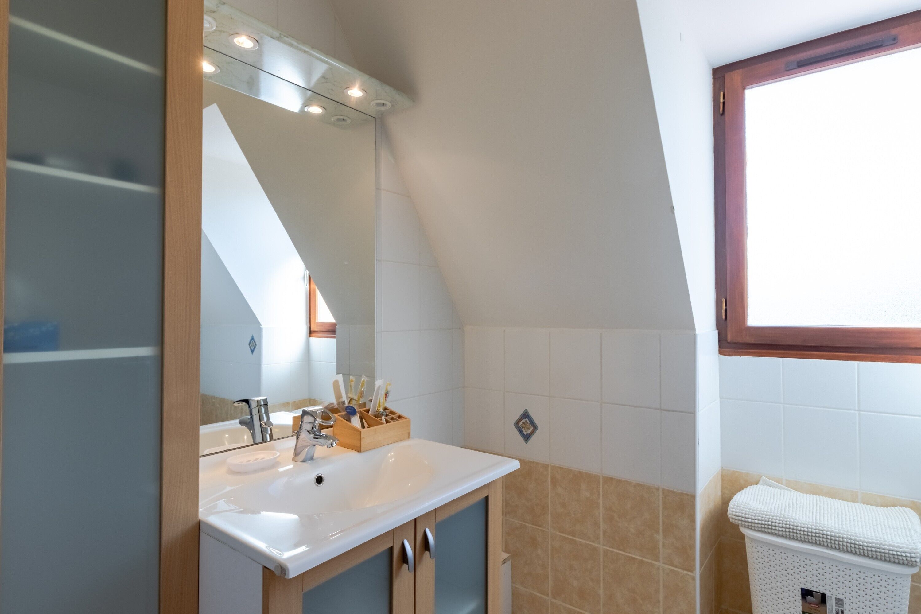Een moderne badkamer met een witte wastafel, houten kastjes, een spiegel met verlichting en een raam.