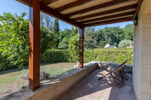 A covered outdoor area with a stone floor, wooden pergola, and a view of a well-maintained garden and distant buildings.