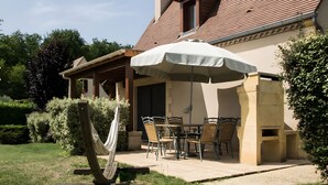Une terrasse couverte, aménagée avec du mobilier d’extérieur, un hamac et offrant une vue sur une maison dont le toit est recouvert de tuiles.