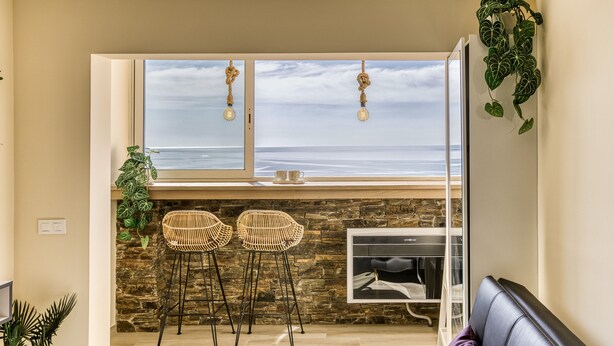Un salón moderno con vista al océano, dos sillones de mimbre, un sofá púrpura y una pared de piedra.