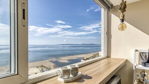 Habitación con amplia ventana que da a una playa, un escritorio de madera y dos tazas sobre un posavasos.