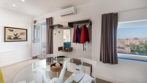 A modern hotel room with a glass dining table, white chairs, a view of the city, and a wardrobe with hanging clothes.