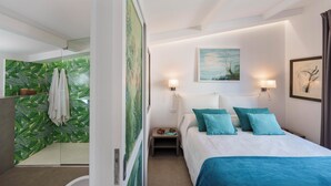 A modern hotel room with a bed, two wall-mounted lamps, a painting, and a shower area with a green leaf-patterned wall.