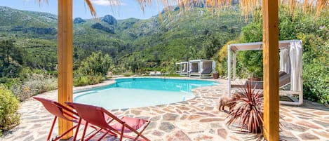 A pool area with a wooden pergola, stone flooring, and a view of mountains.