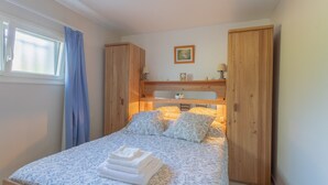 Une chambre à coucher avec un lit, une fenêtre avec des rideaux, une armoire en bois et une étagère avec une lampe et un tableau.
