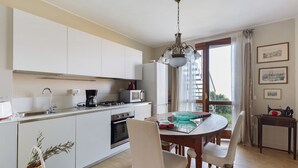 A modern kitchen with white cabinets, a dining table with chairs, a chandelier, and a view through a large window.